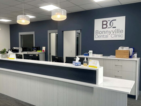A modern dental clinic reception area featuring a white counter, blue walls, and a sign reading "Bonnyville Dental Clinic." The space is well-lit, with stylish light fixtures and organized workstations in the background.