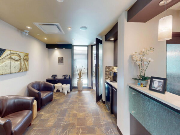 A modern waiting room featuring brown leather chairs, decorative wall art, and a sleek reception desk with plants. Natural light enters through a glass door at the far end, creating a welcoming ambiance.