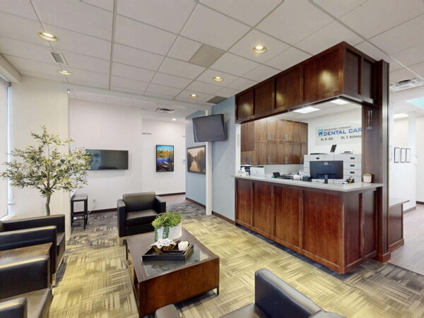 A modern office reception area featuring a wooden front desk, seating arrangements with dark chairs, a decorative plant, and wall-mounted screens. The space is well-lit with large windows and contemporary decor, creating a welcoming environment.