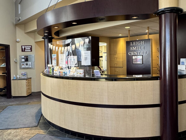 A welcoming dental reception area featuring a curved wooden desk, informative displays, and cheerful lighting. The environment is designed to make patients feel at ease. The 123Dentist Smiley adds a friendly touch to the inviting atmosphere.
