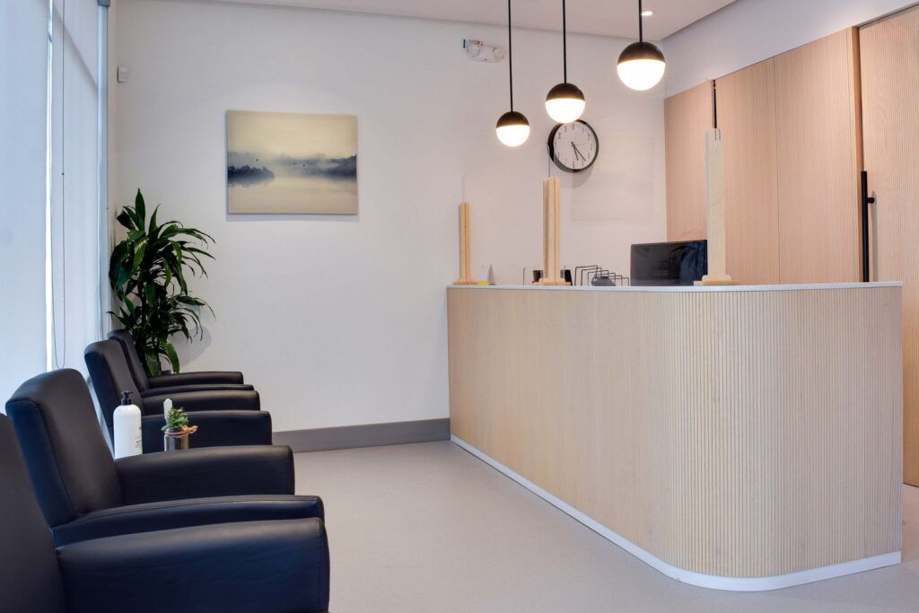 A modern reception area featuring a wooden front desk, black chairs, and a potted plant. The wall has a calming landscape painting, and there are pendant lights and a clock visible. The space is bright and inviting.