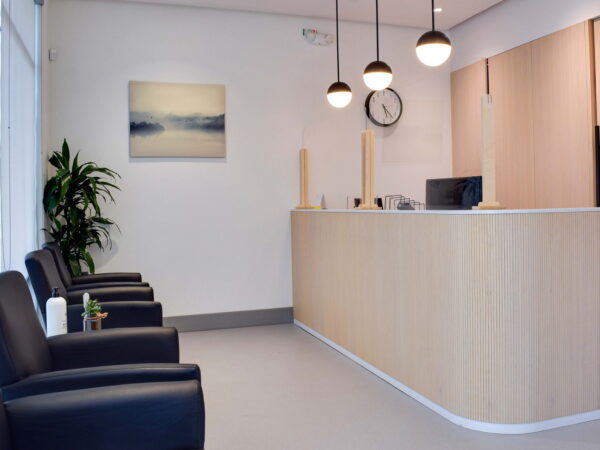 A modern reception area featuring a wooden front desk, black chairs, and a potted plant. The wall has a calming landscape painting, and there are pendant lights and a clock visible. The space is bright and inviting.