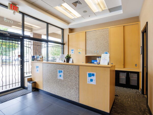 A modern reception area featuring a wooden counter with promotional materials, large windows allowing natural light, and a security gate. The space is neatly organized, with a welcoming atmosphere and a tiled floor.