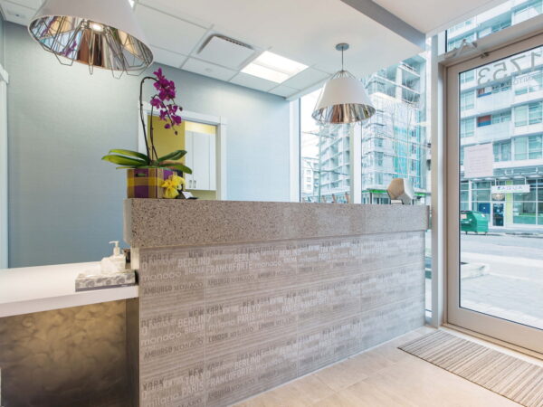 A modern reception area featuring a sleek gray counter with decorative plants and pendant lighting, large windows allowing natural light, and a view of an urban environment outside. The space has a clean, contemporary design.
