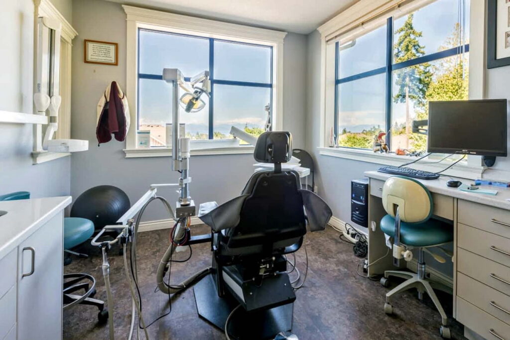 A modern dental office featuring an examination chair, dental equipment, and a computer workstation, illuminated by natural light from large windows. The room is neatly organized and has a calming atmosphere, enhancing patient comfort.