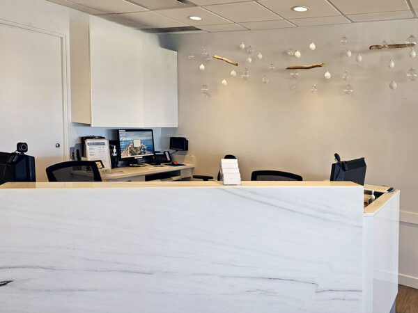 A modern dental office reception area with a white marble front desk, computer monitors, and a bright, inviting atmosphere. Soft lighting hangs from the ceiling, creating a welcoming space for patients. The friendly 123Dentist Smiley adds a cheerful touch to the environment.