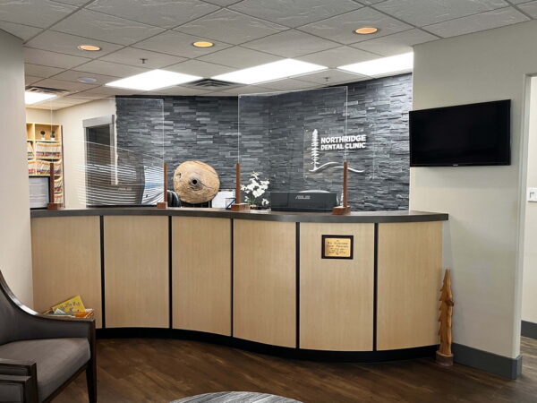 A modern reception area featuring a wooden front desk with a dark stone backdrop. There is a comfortable chair nearby, and the space is well-lit with overhead lights. A television is mounted on the wall, adding to the inviting atmosphere of the office.