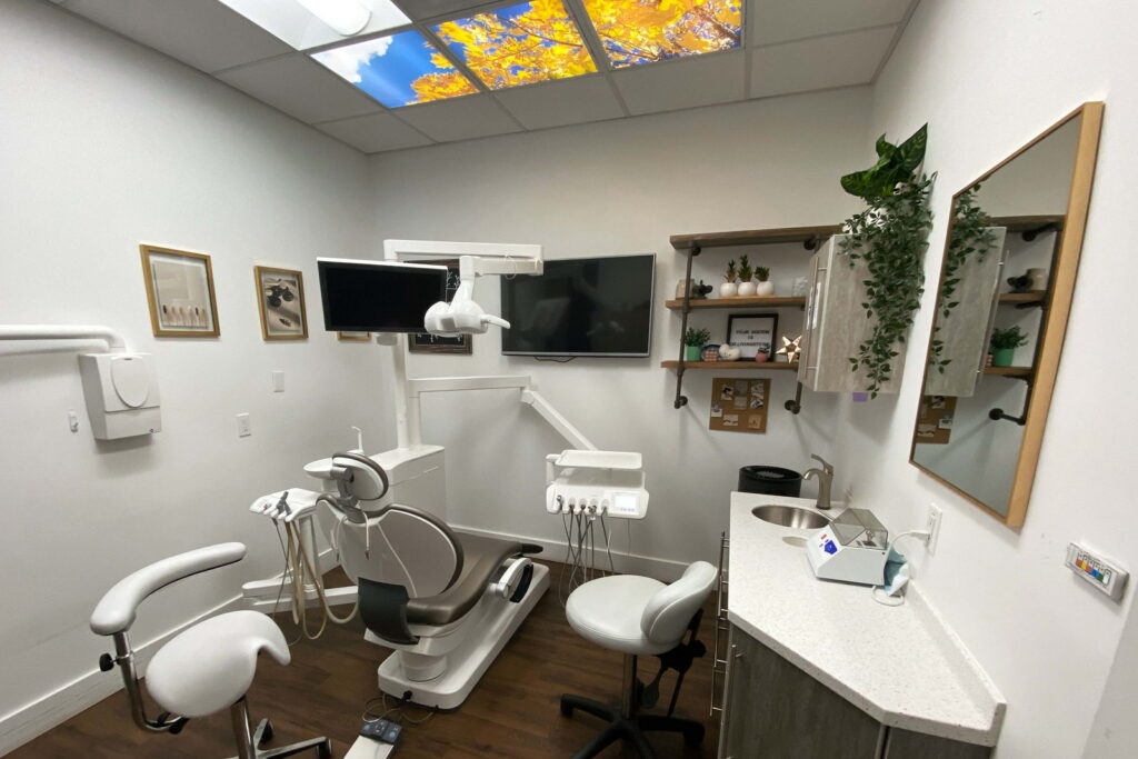 A modern dental office featuring a treatment chair, dental equipment, and a wall-mounted TV. The space is decorated with plants and framed artwork, while a colorful skylight adds natural light to the environment.