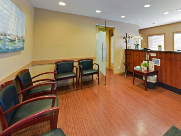 A modern waiting area featuring a reception desk, several chairs, and a decorative painting. The space has warm lighting and wooden flooring, creating a welcoming atmosphere.
