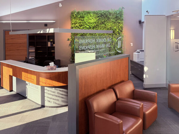 A modern dental reception area featuring stylish wooden desk, cozy brown chairs, and a green living wall. The space is bright and welcoming, designed to create a comfortable atmosphere for patients. 123Dentist Smiley adds a friendly touch to the environment.