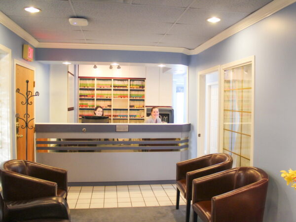 A well-lit reception area featuring a front desk, shelves filled with books, and two people. Comfortable leather chairs are arranged for seating, and there are glass doors leading to another room. The walls are painted blue, creating a welcoming atmosphere.