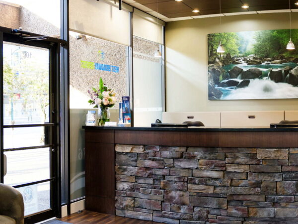 A modern reception area featuring a stone counter, brightly lit interior, and a large nature-themed artwork of a stream. There are flowers on the counter and glass doors leading outside.