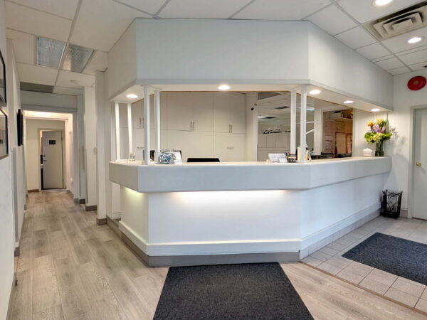 A well-lit reception area with a white counter and floral arrangements. The space features gray flooring, minimal decor, and a hallway leading to private rooms.