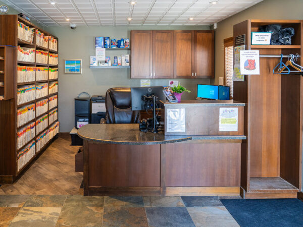 A dental office reception area with wooden furniture, file storage, and a welcoming atmosphere. The front desk is organized with plants and signage, inviting patients into a clean and professional environment.