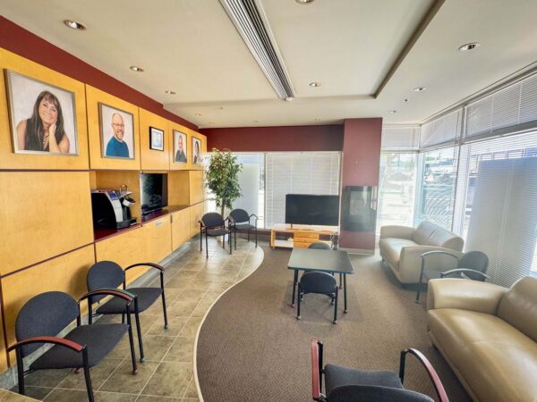 A modern waiting area featuring comfortable seating, a coffee machine, and a television. The walls display framed portraits of individuals, and large windows allow natural light to brighten the space.