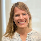 A smiling woman with long, light brown hair and a friendly expression is wearing a patterned blouse. The background is soft and neutral, enhancing her welcoming demeanor.