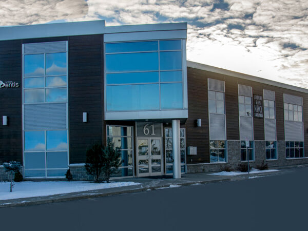 A modern dental clinic building with large windows, dark wood siding, and a prominent address number "61" on the entrance. The sky above is partly cloudy.