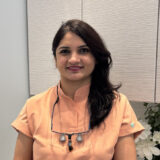 A smiling dental professional in a peach-colored uniform stands against a neutral background, showcasing a friendly and welcoming demeanor.