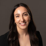 A smiling woman with long, dark hair and hoop earrings poses against a neutral background. She wears a black top and exudes warmth and friendliness.
