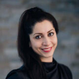 A smiling woman with long dark hair is wearing a black top against a softly blurred background. Her warm expression conveys friendliness and approachability, making her seem welcoming and confident.