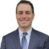 A smiling man in a suit, wearing a white shirt and tie. He has short dark hair and is standing against a plain background, exuding a professional and approachable demeanor.