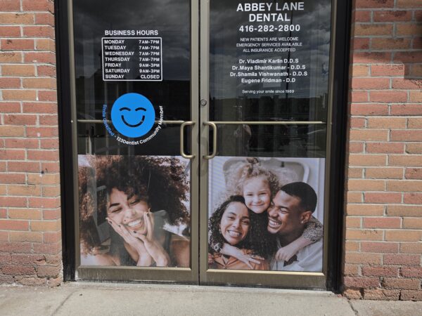 Dental clinic entrance with business hours and contact info on the door. Joyful family photo and the friendly 123Dentist Smiley adorn the glass, set against a brick wall.
