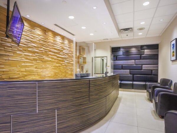 A modern reception area featuring a curved wooden front desk, stylish chairs, and a wall with decorative panels. Soft lighting enhances the welcoming atmosphere, complemented by a wall-mounted TV and framed artwork.