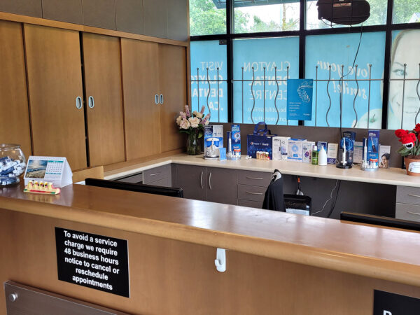 A reception area featuring a wooden counter, a glass bowl of candies, various brochures, and decorative flowers. Large windows allow natural light, and a sign is visible on the counter.