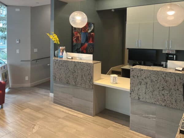 A modern dental office reception area featuring a sleek counter with a granite finish, a cozy red chair, and bright lighting. On the wall, colorful artwork adds vibrancy to the space, creating a welcoming environment. The friendly 123Dentist Smiley is nearby, ready to greet visitors.