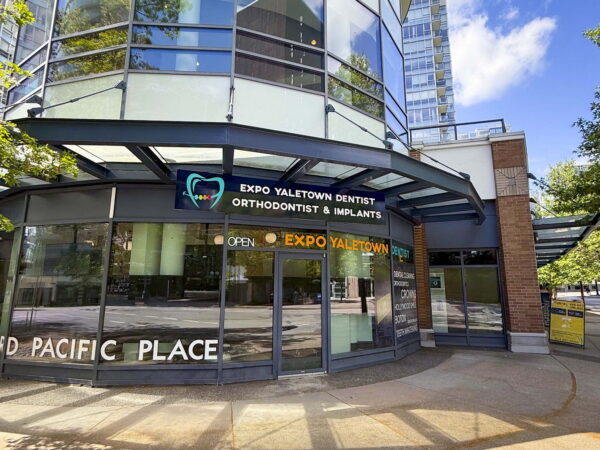 A modern dental office exterior featuring large glass windows and signage for "Expo Yaletown Dentist, Orthodontist & Implants" at Concord Pacific Place. The building is surrounded by trees and bright blue sky, showcasing a welcoming urban environment.
