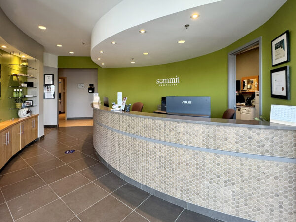 A modern dental reception area featuring a curved front desk, green walls, and warm lighting. The space includes a waiting area with chairs and various dental materials on display, providing a welcoming atmosphere for patients.