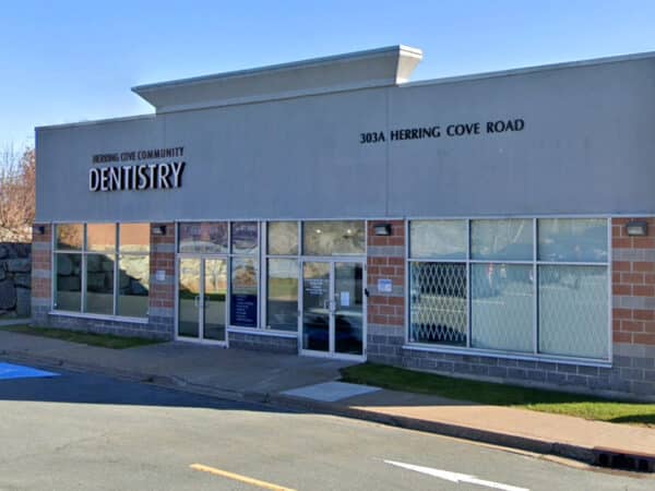 A modern dental clinic building with large windows and a sign that reads "Dentistry" prominently displayed. The entrance includes sliding glass doors and is located on a road named "Heritage Cove Road," surrounded by a well-maintained parking area.