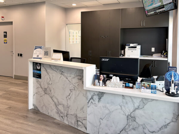 A modern dental office reception area featuring a marble front desk, a computer station, and various dental supplies. The space is well-lit and organized, creating a welcoming atmosphere for patients. 123Dentist Smiley is nearby, adding a friendly touch to the environment.