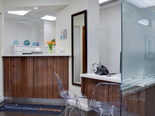 A modern dental office reception area featuring a sleek wooden front desk, clear acrylic chairs, a glass partition, and vibrant floral decor. The space is tidy and well-lit, promoting a welcoming atmosphere for patients.
