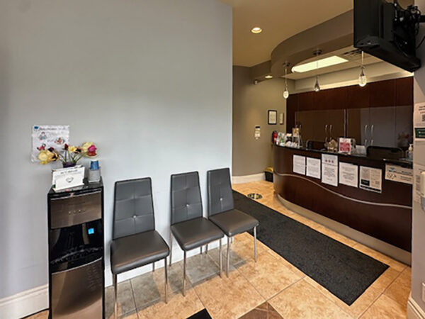 A dental office waiting area featuring three gray chairs, a water cooler, and a reception desk in the background. The space is well-lit and organized, creating a welcoming environment for patients.