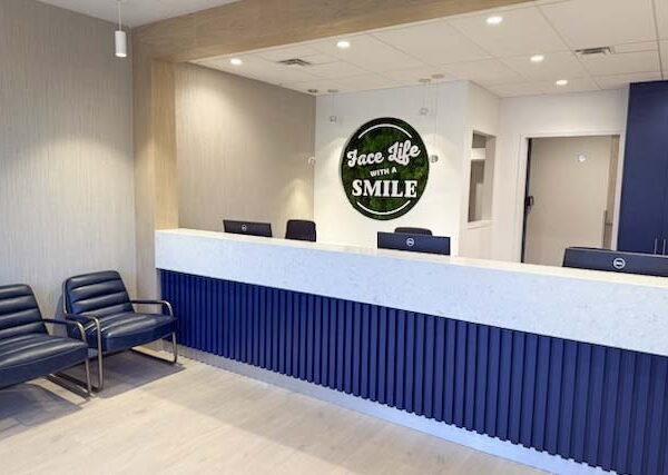 A modern reception area featuring a sleek front desk with a "Face Life Smile" sign, blue accent walls, and two black chairs for waiting clients.