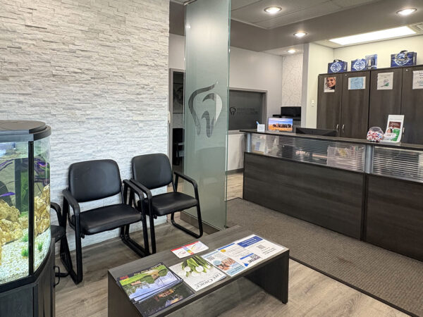 A dental office interior featuring a reception area with a counter, two black chairs, and a coffee table displaying magazines. There is an aquarium visible, providing a calming environment for patients. The 123Dentist Smiley can be seen welcoming visitors.