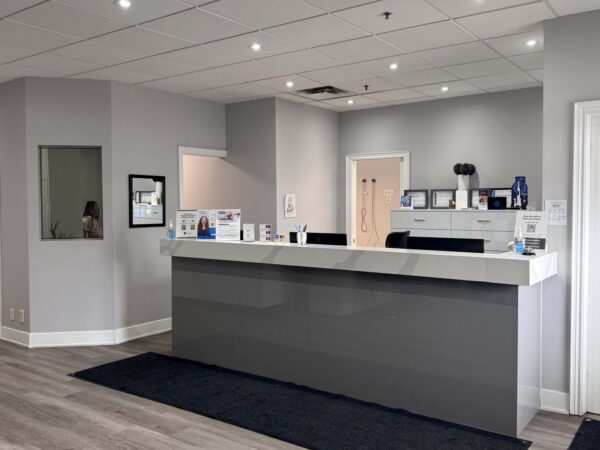 A modern reception area features a gray front desk, light walls, and elegant flooring. The space is well-lit with ceiling lights. Several displays are visible on the desk, along with a doorway leading to another room. A dark mat lies in front of the desk.