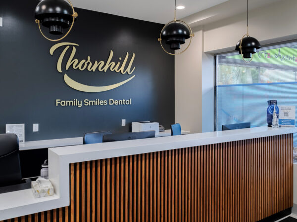 L'image montre la zone d'accueil de Thornhill Family Smiles Dental, présentant un design moderne avec un comptoir blanc, des accents en bois et un éclairage suspendu élégant. Il y a des documents d'information sur le comptoir et une vue de l'extérieur à travers de grandes fenêtres.