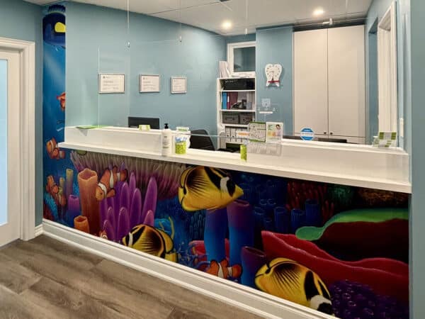 A bright dental office featuring a colorful fish mural on the reception wall, creating a cheerful atmosphere. The front desk is modern and welcoming, designed to make patients feel at ease. The 123Dentist Smiley adds a friendly touch to the environment.