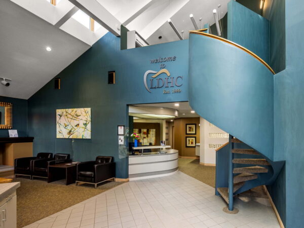The image shows a modern reception area with a spiral staircase. The walls are painted blue, adorned with artwork and a logo. Comfortable seating is arranged near the reception desk, creating an inviting atmosphere.