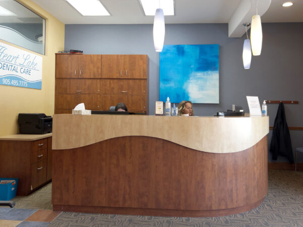 A welcoming dental office reception area featuring a wooden front desk with a curved design, modern light fixtures, and a vibrant blue abstract painting in the background. The space is tidy and inviting, ready to serve patients. 123Dentist Smiley adds a friendly touch to the environment.