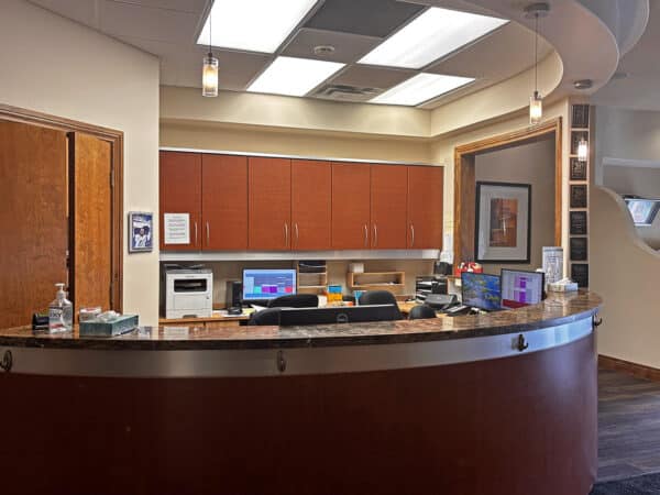 A modern dental office reception with a curved desk, wooden cabinetry, and digital screens. Bright lighting highlights the inviting atmosphere, creating a friendly environment for patients awaiting their appointments.
