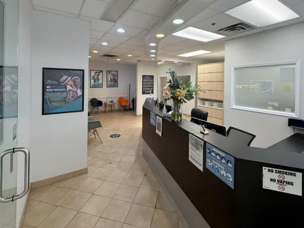 A modern dental office reception area featuring a dark wooden front desk, plants, and seating. Promotional posters adorn the walls, while bright lighting creates a welcoming atmosphere. The friendly 123Dentist Smiley adds a cheerful touch to the space.