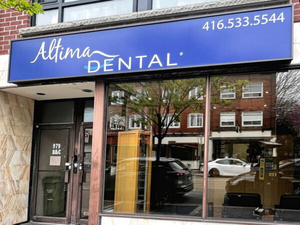 A dental clinic storefront featuring a blue sign that reads "Altima DENTAL" along with a phone number. The entrance is framed by glass windows showing a view of the street outside.