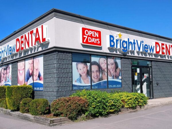 A dental clinic building featuring the sign "BrightView DENTAL" prominently displayed. The exterior includes large windows with portraits of smiling patients and a banner indicating it is open seven days a week. Lush greenery surrounds the entrance.
