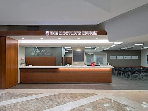 A modern doctor's office reception area featuring a wooden desk with the sign "The Doctor's Office," behind which is a waiting area with chairs. The space is well-lit and has a clean, professional appearance.