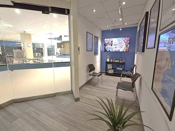 A modern waiting area featuring a sleek design. It includes chairs, artwork on the walls, and a television screen displaying images. Natural light fills the space through large windows, creating a welcoming atmosphere. A potted plant adds a touch of greenery.