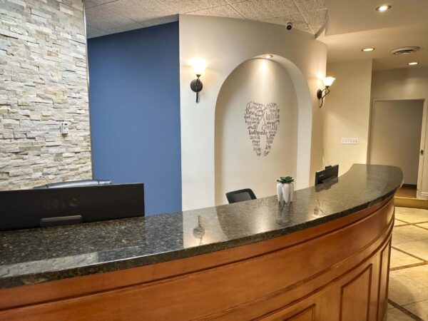A modern reception area featuring a curved wooden desk with a dark countertop, a stone accent wall, and a blue wall with a heart-shaped art piece. The space is well-lit and includes potted plants for decoration.
