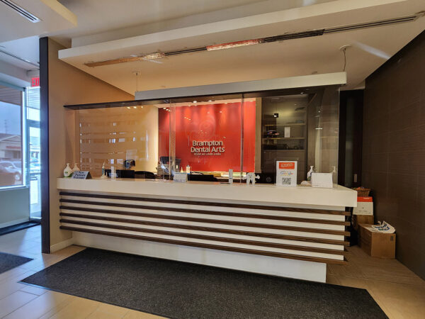 A modern reception area featuring a sleek front desk with a wooden design, glass panels, and an orange accent wall. The space is well-lit and includes entry mats, creating a welcoming atmosphere for visitors.